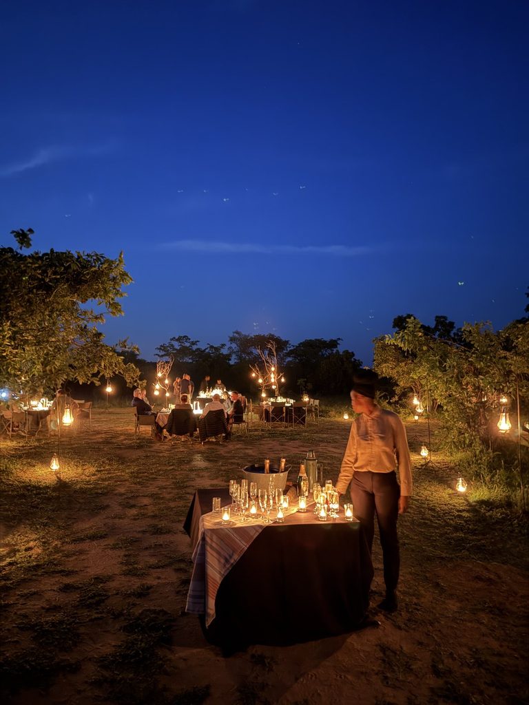 Lantern-lit outdoor bush dinner setup at a Sabi Sands safari lodge, with a server standing by a table of glasses and champagne under a twilight sky
